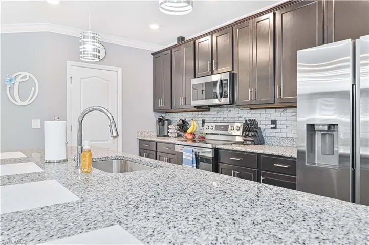 Kitchen with dark brown cabinetry, appliances with stainless steel finishes, decorative backsplash, light stone counters, and crown molding