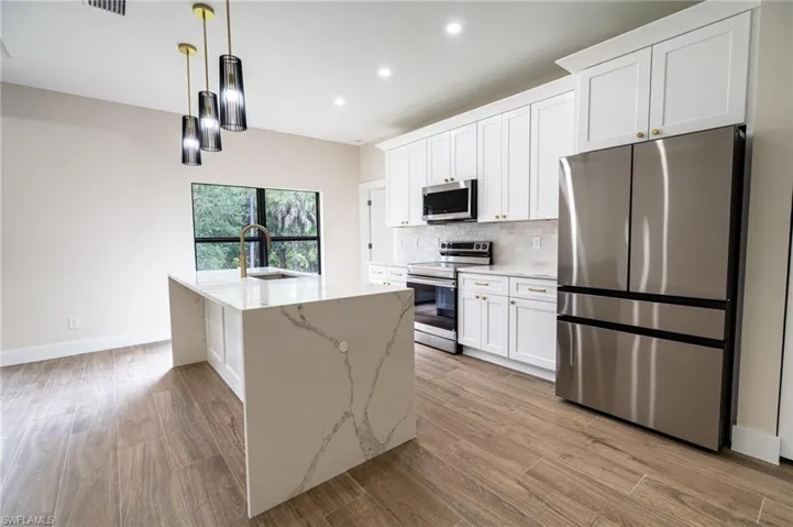 Kitchen featuring a waterfall island with a gold-finish faucet, pendant lighting, white shaker cabinetry, stainless steel appliances, and wood-finish flooring