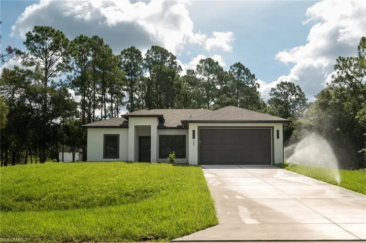 Modern single-story residence featuring a two-car garage, concrete driveway, front lawn, and mature trees framing the property