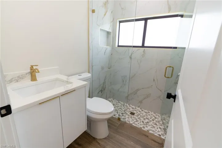 Modern bathroom featuring a white vanity with gold hardware, a glass-enclosed shower with pebble flooring, full-height wall tiling, and wood-finish flooring