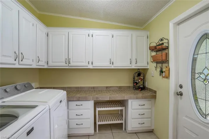 Huge laundry room features white upper cabinetry, tiled countertop,  built-in lower cabinetry with drawers, and a white entry door with decorative glass inlay.