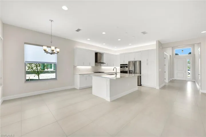 Kitchen featuring white cabinetry, wall chimney exhaust hood, stainless steel appliances, a center island with sink, and view of front door/foyer as well as Breakfast Area with chandelier