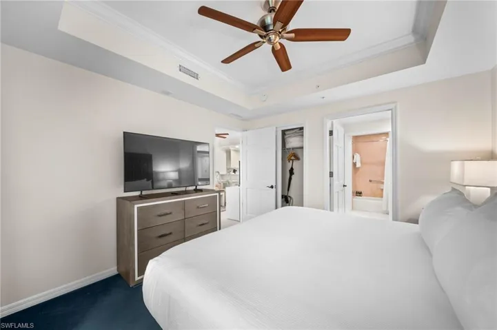Bedroom featuring ornamental molding, a tray ceiling, a walk in closet, dark colored carpet, and a ceiling fan