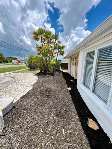 Landscaped exterior featuring a textured paver driveway, dark mulch beds, mature palm trees, white siding, and prominent windows with horizontal blinds