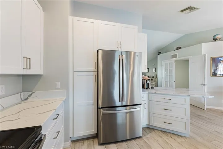 Kitchen featuring white Shaker-style cabinetry, stainless steel French-door refrigerator, stone-look countertops with veining, and wood-finish flooring under a vaulted ceiling