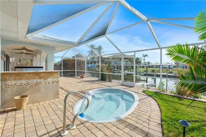 View of swimming pool featuring glass enclosure, a water view, ceiling fan, a patio area, and exterior kitchen