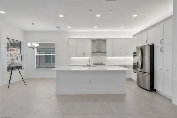 Kitchen featuring wall chimney range hood,  stainless steel fridge, a kitchen island with sink, and light tile floors