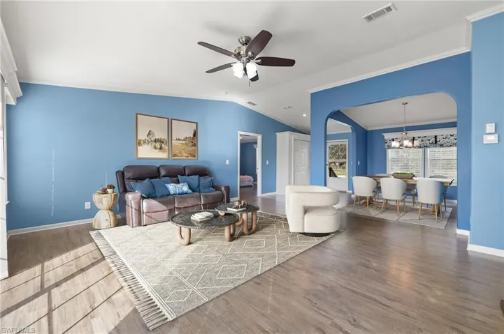 Living room with ceiling fan with notable chandelier, hardwood / wood-style flooring, vaulted ceiling, and crown molding, partially staged virtually