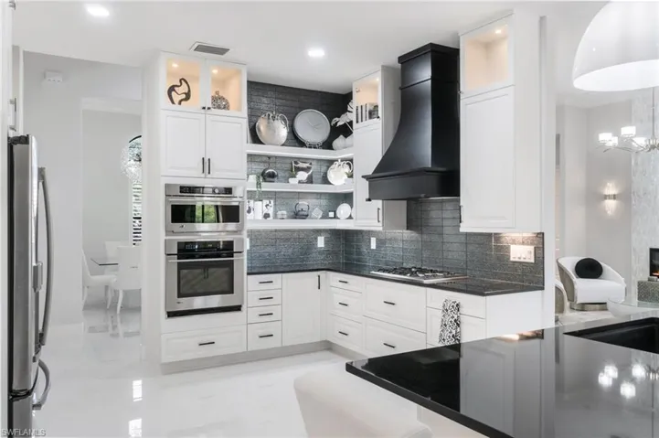 Modern kitchen featuring white cabinetry with black hardware, stainless steel built-in appliances, a black range hood, dark countertops, and a dark tile backsplash