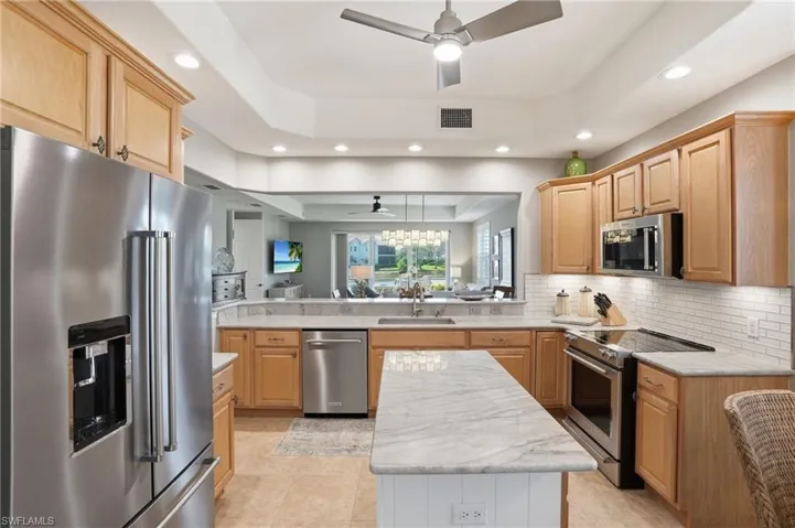 Kitchen with stainless steel appliances, a raised ceiling, ceiling fan, light wood finish cabinetry, and recessed lighting
