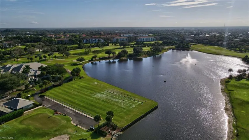 Expansive golf course featuring a large water hazard with a central fountain, manicured fairways, mature trees, and nearby residential buildings