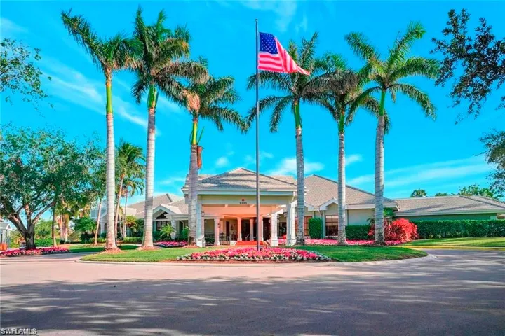Circular driveway entrance with a central landscaped island featuring a flagpole, bordered by palm trees and vibrant flowering plants