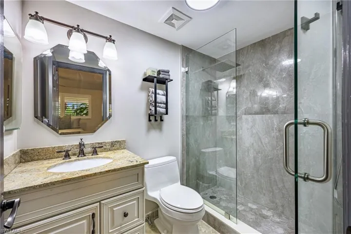 Bathroom featuring a glass-enclosed shower with gray stone-finish wall tile, a white vanity with a light-toned countertop, an octagonal mirror, a three-light vanity fixture, and a white toilet