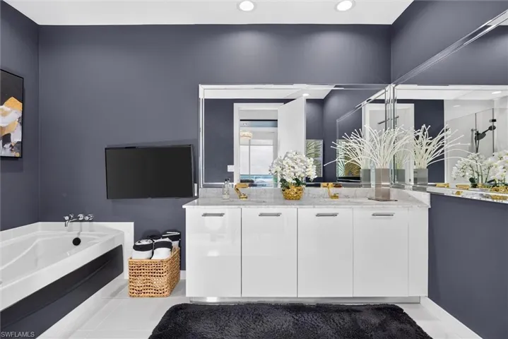 Dual vanity with a stone countertop, wall-mounted television, built-in bathtub, recessed lighting, and wall-mounted mirrors
