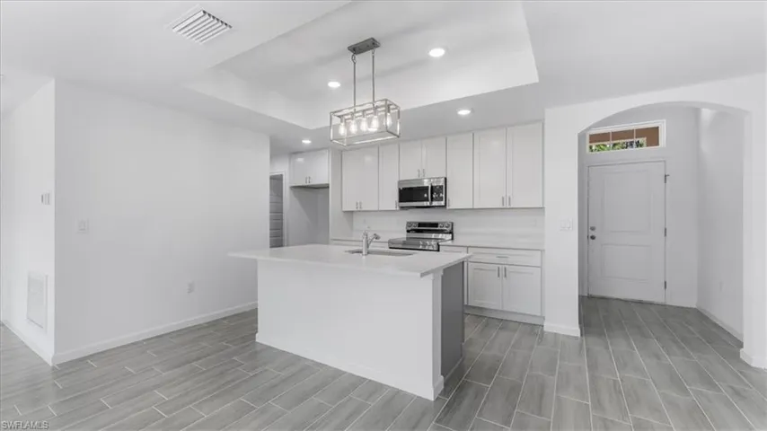 Kitchen with white cabinets, stainless steel appliances, a raised ceiling, a center island with sink, and hanging light fixtures