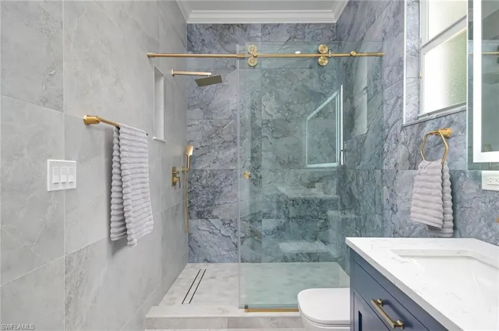 Primary Bathroom featuring an enclosed shower with custom doors, tile walls, toilet, double vanity, and ornamental molding