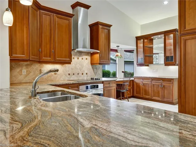 Kitchen featuring decorative light fixtures, brown cabinetry, wall chimney exhaust hood, and light stone counters