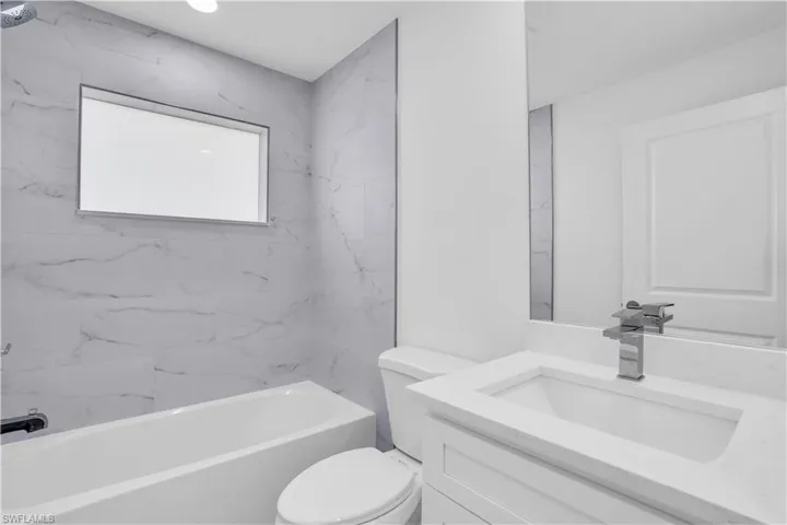 Bathroom featuring a white bathtub, marble-patterned wall tile, a window, a white vanity with an integrated sink, and a chrome faucet