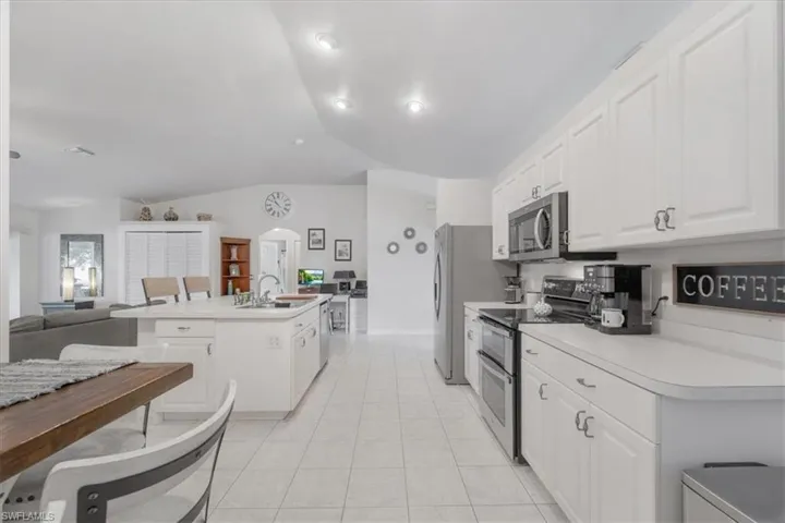 Kitchen with appliances with stainless steel finishes, white cabinets, vaulted ceiling, light tile patterned floors, and an island with sink