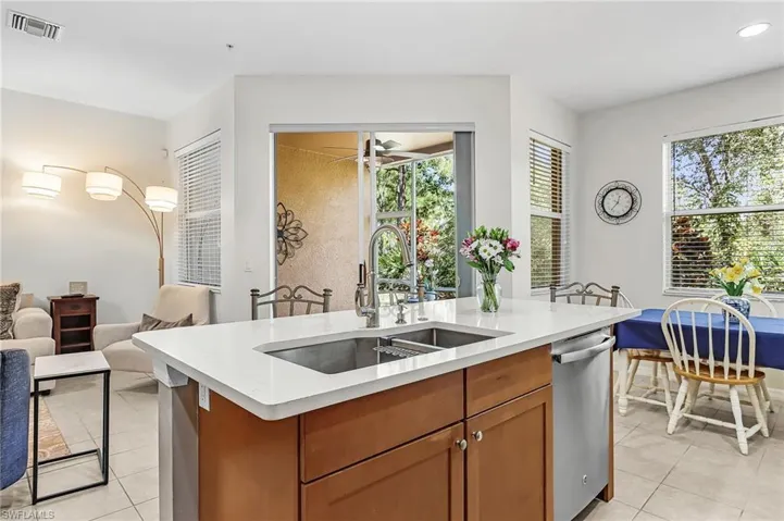 Kitchen featuring wood finish cabinetry, open floor plan, an island with sink, stainless steel dishwasher, and light stone countertops