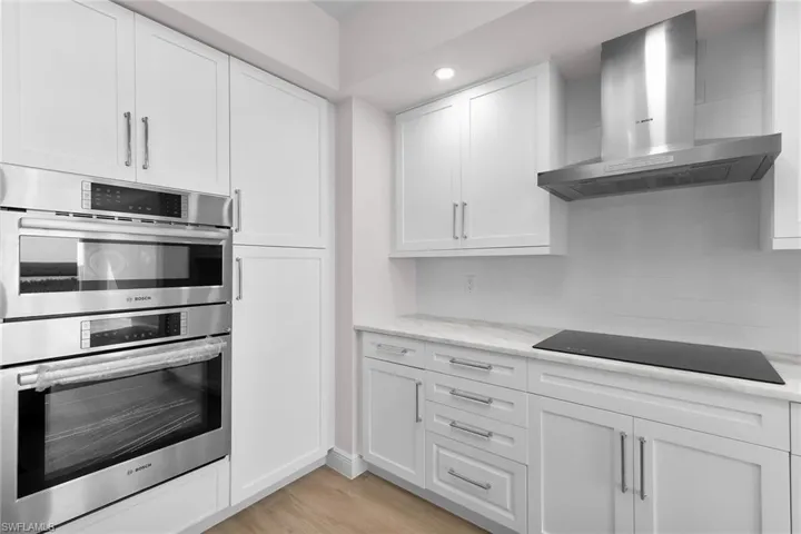 Kitchen featuring wall chimney exhaust hood, double oven, white cabinets, light wood-style floors, and black electric cooktop