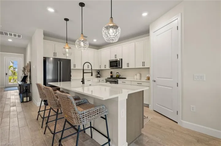 Kitchen featuring light wood-type tile flooring, stainless steel appliances, a center island with sink, white cabinetry, and hanging light fixtures