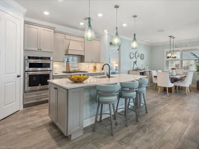 Kitchen featuring an island with sink, double oven, custom exhaust hood, hanging light fixtures, and decorative backsplash