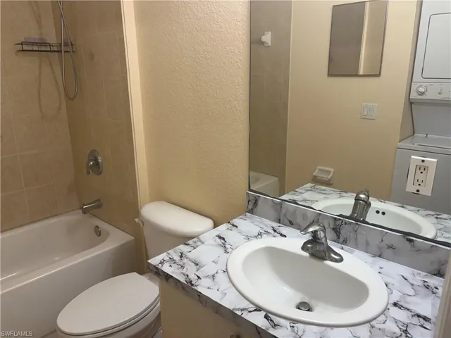 Bathroom featuring a tub/shower combination with tiled surround, a toilet, vanity with a white oval sink, and a stacked washer and dryer unit