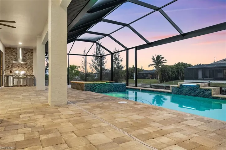Pool at dusk with glass enclosure, a patio area, pool water feature, area for grilling, and an in ground hot tub