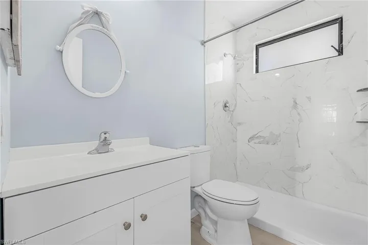 Bathroom featuring a white vanity with a solid surface countertop, an oval mirror, a white toilet, and a bathtub with white marble-look wall tiling