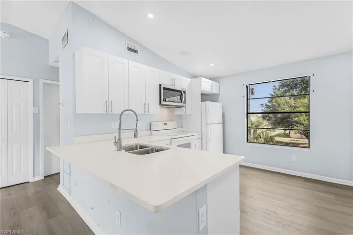 Open-concept kitchen featuring white shaker cabinetry, light-toned countertops, stainless steel sink with gooseneck faucet, and wood-finish flooring