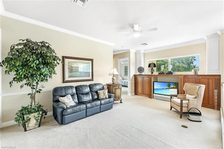 Carpeted room featuring crown molding, built-in cabinetry with wood-finish doors, a window overlooking exterior greenery, and a ceiling fan