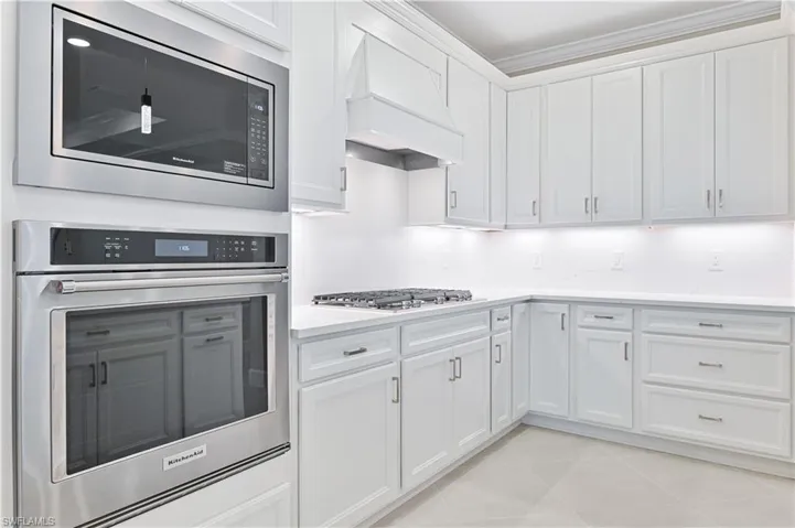 Kitchen featuring stainless steel appliances, custom range hood, white cabinetry, crown molding, and light stone counters
