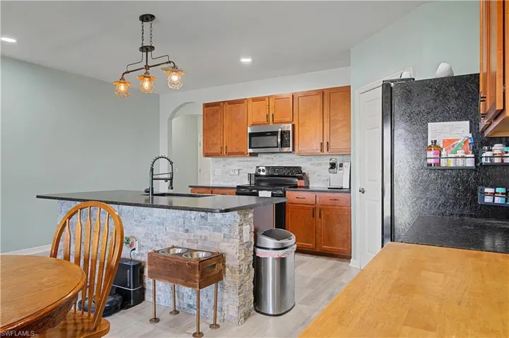 Kitchen with pendant lighting, arched walkways, brown cabinets, appliances with stainless steel finishes, and an island with sink