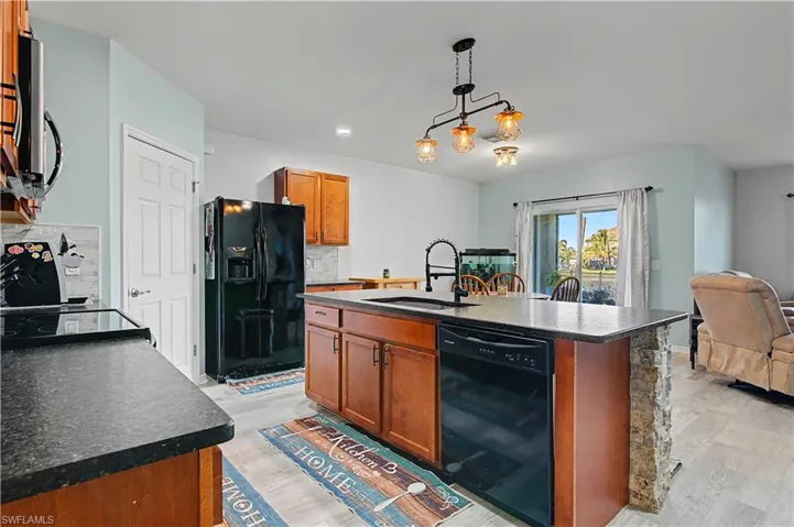 Kitchen featuring brown cabinetry, a kitchen island with sink, black appliances, dark countertops, and decorative backsplash