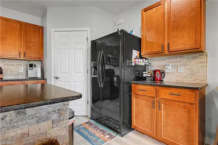 Kitchen featuring decorative backsplash, brown cabinetry, black fridge with ice dispenser, dark stone countertops, and light wood-style flooring