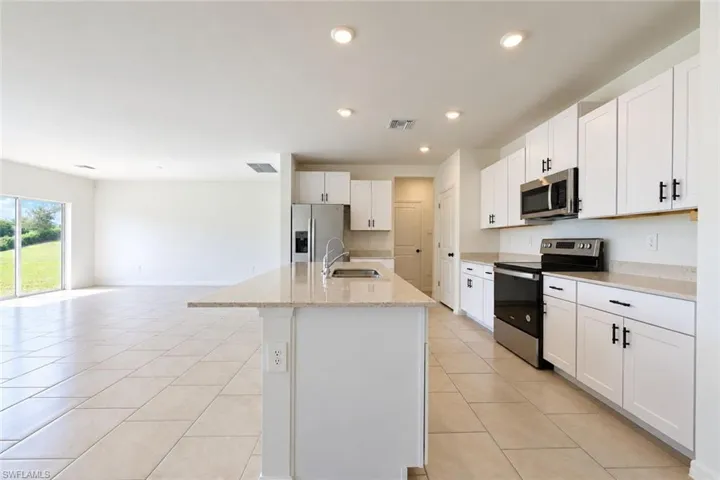 Kitchen featuring white shaker-style cabinetry, stone-composite countertops, stainless steel appliances, a center island with an integrated sink, and large-format ceramic tile flooring