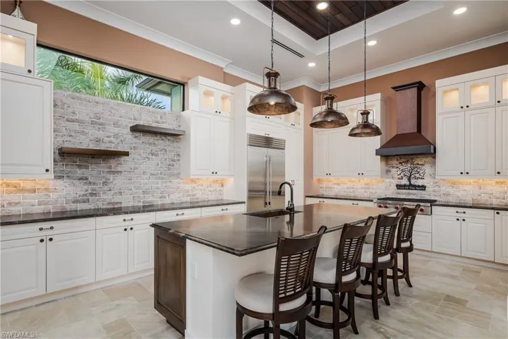 Gourmet kitchen featuring a large central island with seating, white shaker-style cabinetry, a distressed brick backsplash, and undermount lighting