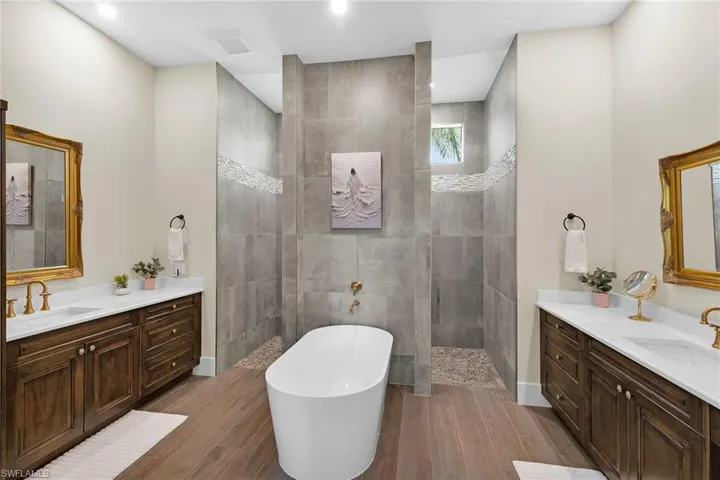 Freestanding soaking tub with brushed gold fixtures, flanked by dual wood-finish vanities featuring white countertops and undermount sinks