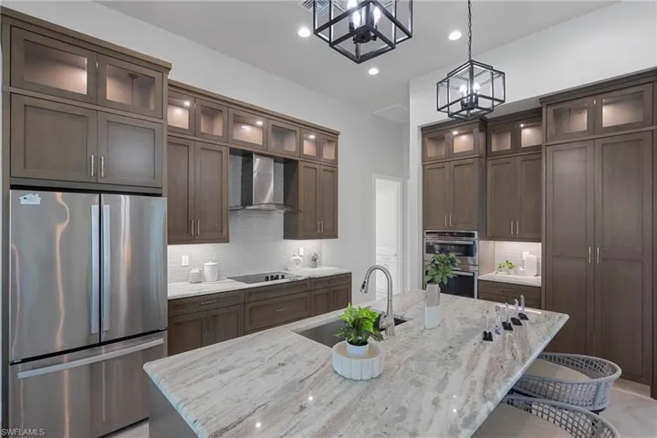 Kitchen with wall chimney range hood, sink, stainless steel appliances, light stone countertops, and an island with sink