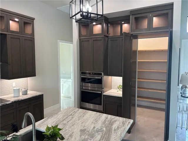 Kitchen featuring backsplash, hanging light fixtures, double oven, dark brown cabinets, and light stone counters