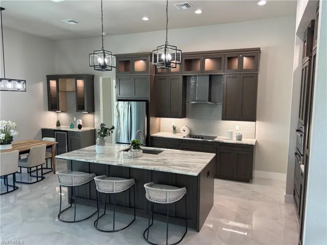 Kitchen with stainless steel refrigerator, wall chimney range hood, decorative backsplash, light stone counters, and a kitchen island with sink