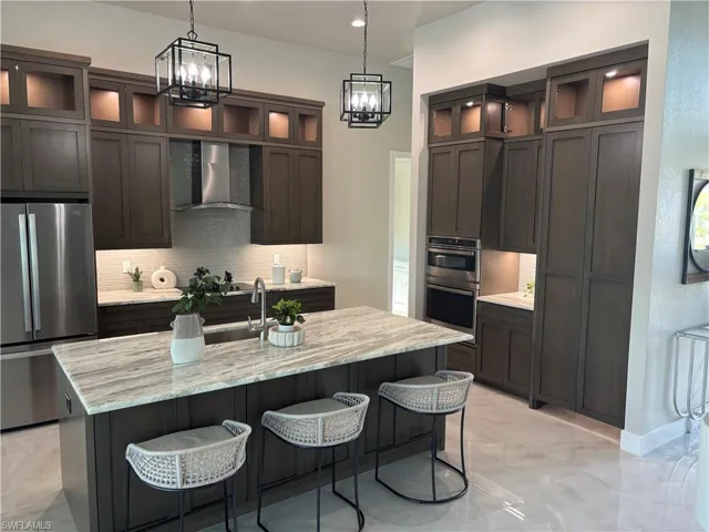 Kitchen featuring light stone countertops, tasteful backsplash, stainless steel appliances, a kitchen island with sink, and wall chimney range hood