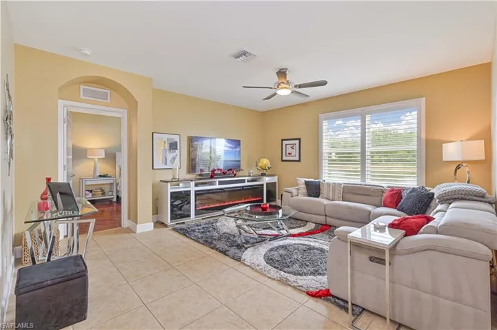 Spacious living area featuring neutral tile flooring, a ceiling fan with integrated lighting, and a large window with white plantation shutters