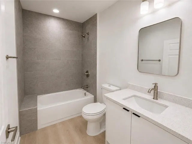Full bathroom featuring a white bathtub with a tiled shower surround and integrated bench, wood-finish flooring, a white vanity with a speckled countertop and integrated sink, a brushed nickel faucet, and a contemporary wall-mounted mirror