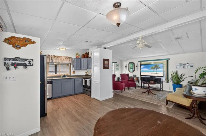 Open-concept living area featuring wood-finish flooring, galley-style kitchen with blue cabinetry and white countertops, ceiling fans, and multiple windows with natural light throughout the space