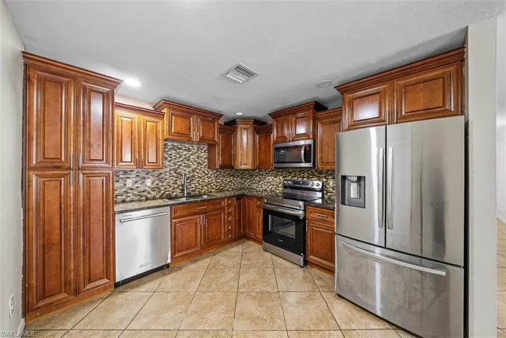 Kitchen featuring wood cabinetry, stainless steel appliances, mosaic tile backsplash, dark countertops, and tile flooring