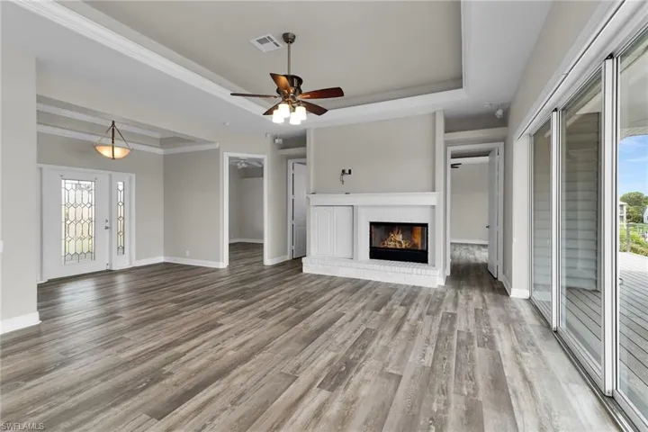Unfurnished living room featuring a raised ceiling, wood finished floors, a brick fireplace, ceiling fan, and ornamental molding