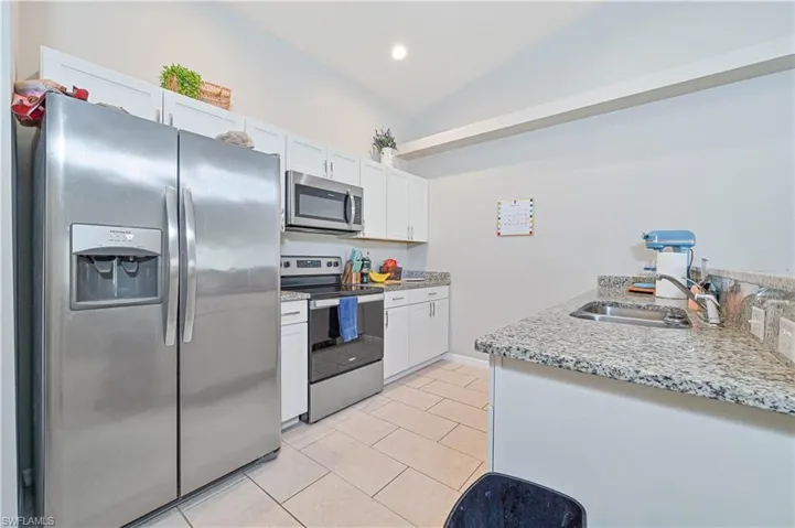 Kitchen featuring stainless steel appliances, vaulted ceiling, light tile patterned floors, white cabinets, and light stone counters