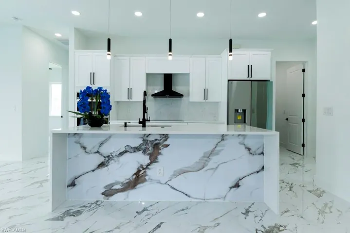 Kitchen with stainless steel refrigerator with ice dispenser, wall chimney range hood, a kitchen island with sink, recessed lighting, and white cabinetry